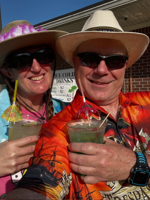 Two people smiling and posing for a selfie, each holding a colorful drink with umbrellas, wearing sun hats and sunglasses, with a brick wall and a drinks sign in the background.