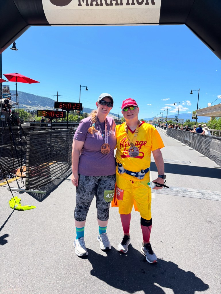 Two runners celebrating at the finish line of the Missoula Marathon, wearing medals and colorful outfits, with a clear blue sky in the background.