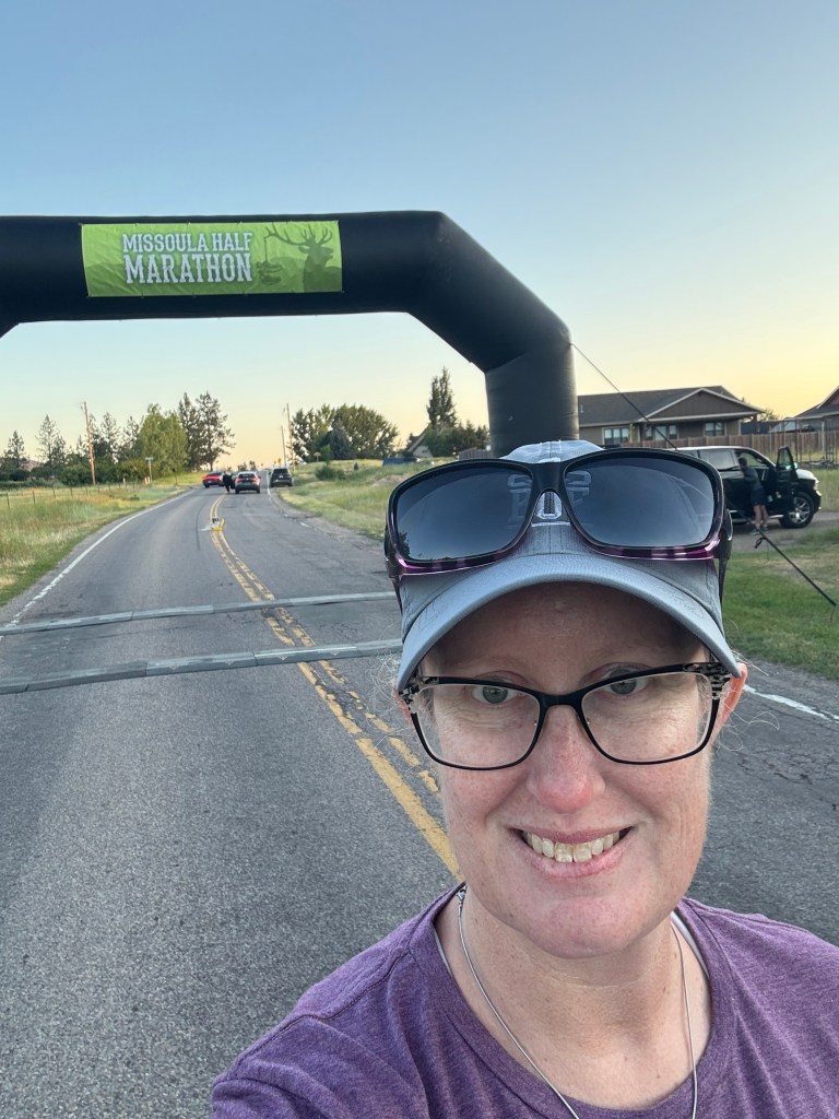 A person poses for a selfie in front of the finish line archway of the Missoula Half Marathon, with trees and vehicles in the background.
