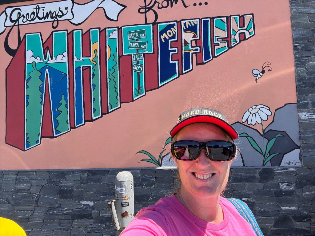 A person smiling in front of a colorful mural that says 'Greetings from Whitefish, Montana', featuring trees and a sunny background.
