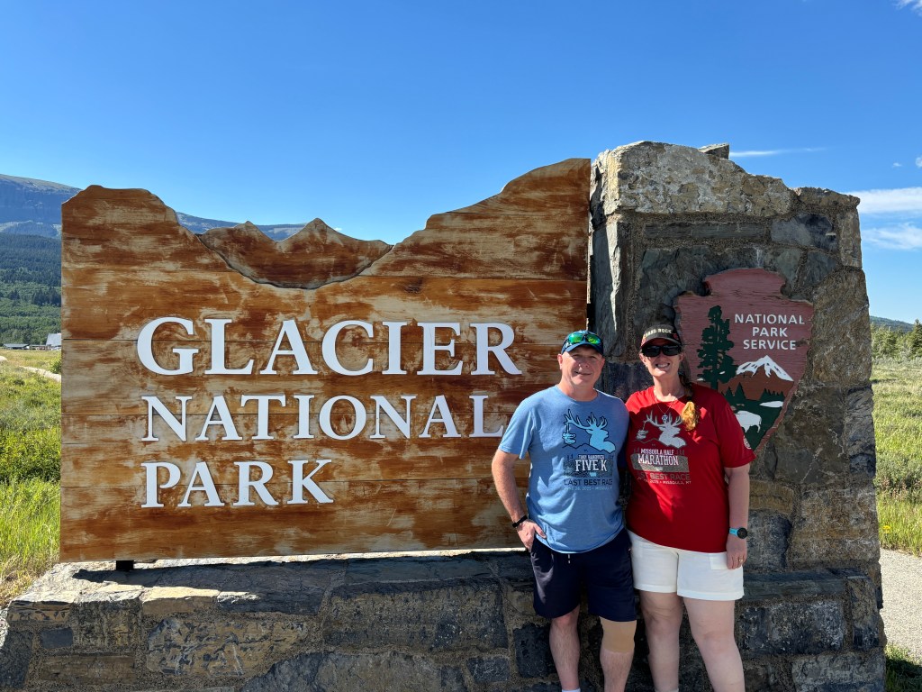 A couple standing in front of the Glacier National Park entrance sign, enjoying their visit to the park.