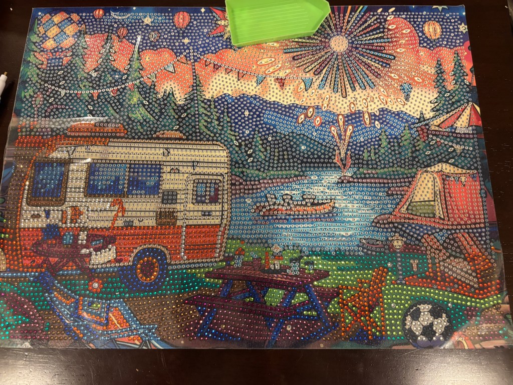 A colorful diamond painting featuring a vintage camper by a lake, surrounded by trees and tents, with fireworks in the sky above.