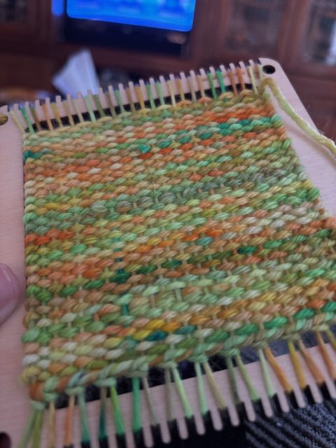 Close-up of a woven coaster on a loom, featuring vibrant shades of green, orange, and yellow yarn.