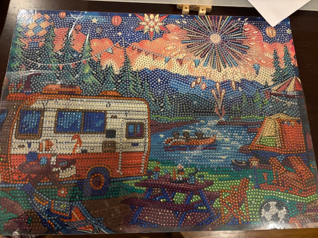A vibrant diamond painting depicting a scenic campsite with a colorful sunset, an RV, a campfire, and boats on a lake surrounded by trees.