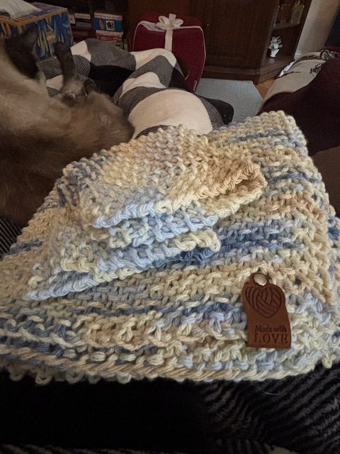 A cozy knitted kitchen towel and two matching dishcloths arranged on a couch, featuring a wooden tag that says 'Made with Love' and a soft background with a pet visible.