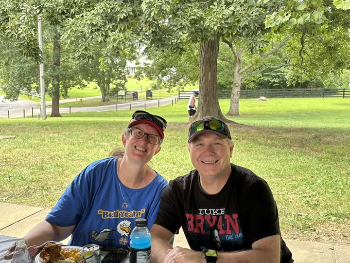 Two smiling individuals sitting at a picnic table outdoors in a park, surrounded by greenery.
