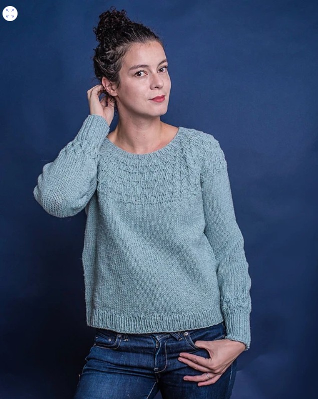 A woman wearing a light blue knitted sweater with a textured yoke, posing against a dark blue background.