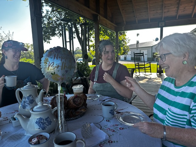A cheerful outdoor birthday celebration featuring three women enjoying tea and pastries at a table decorated with a 'Happy Birthday' balloon.