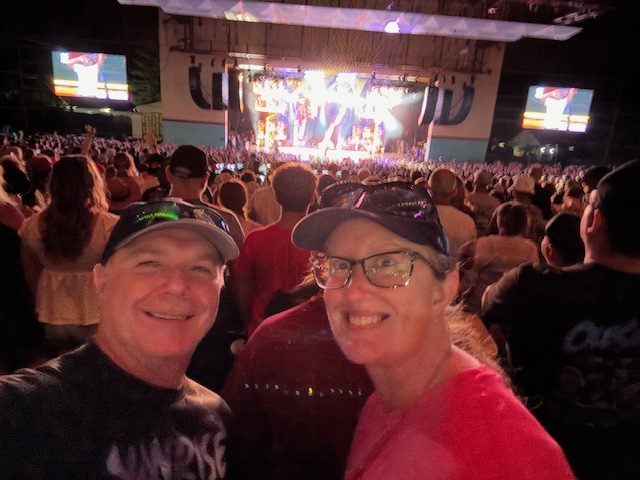 A selfie of two people smiling at a concert, with a large crowd in the background and a brightly lit stage featuring a performer.