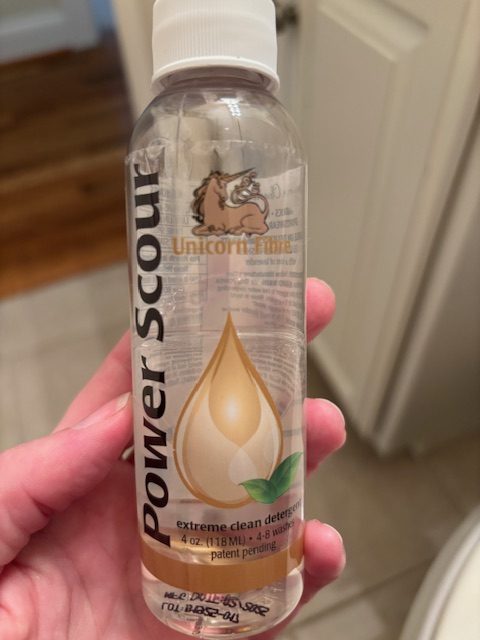 A hand holding a small bottle of Unicorn Power Scour detergent, labeled as an extreme clean product for fleece, with a clear liquid inside.