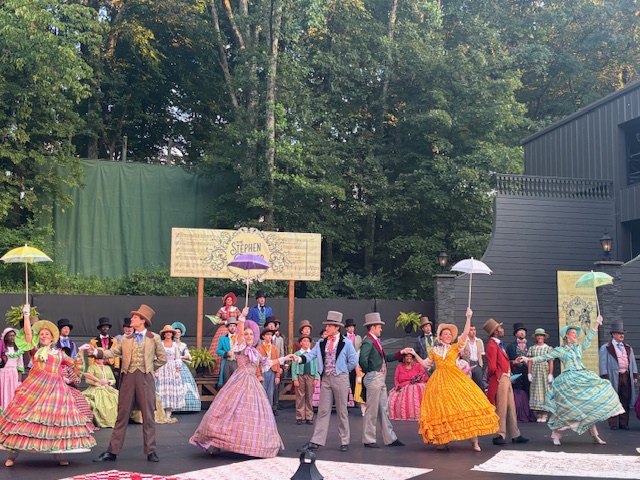 A vibrant outdoor theater performance featuring actors in colorful period costumes, dancing and posing with umbrellas, set against a backdrop of trees.