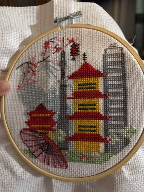 A completed cross-stitch project featuring a Japanese pagoda, cherry blossoms, an umbrella, and a city skyline, displayed in a wooden embroidery hoop.
