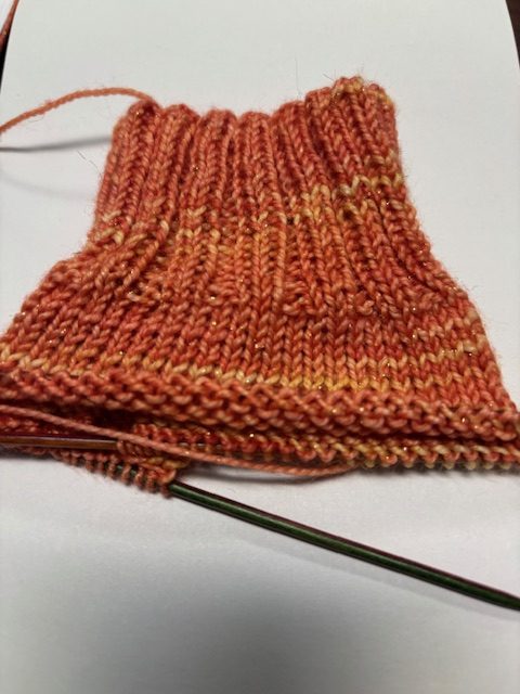 A close-up image of a partially knitted sock in shades of orange with a ribbed pattern, showing progress on knitting with a pair of needles.