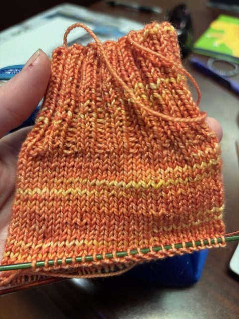 A close-up of a partially knitted orange sock, featuring varying shades of orange and yellow yarn.