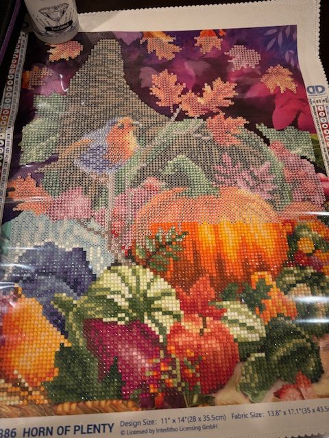 A partially completed diamond painting featuring pumpkins, autumn leaves, and a bird, laid out on a crafting table.