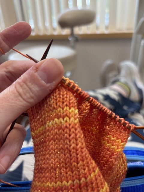 Close-up of a hand holding knitting needles with orange and yellow yarn, while sitting in a dentist's office.
