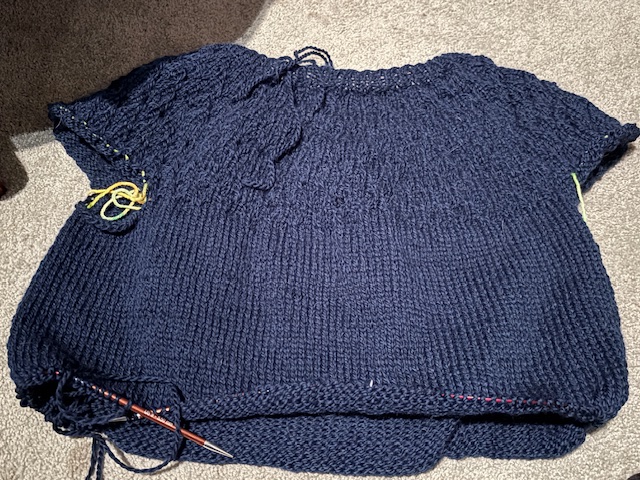 A progress shot of a dark navy knitted sweater laid flat on a light-colored carpet, showing the body and part of the sleeves, with knitting needles and colorful yarn markers attached.
