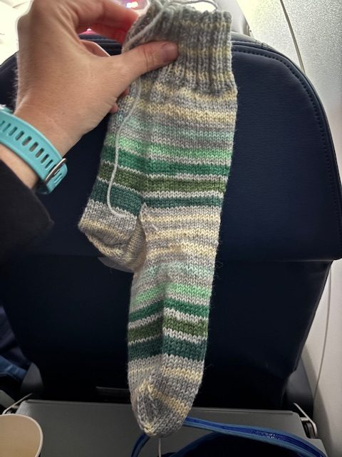 A hand holding a completed striped sock, showcasing green, gray, and yellow hues, with an airplane seat in the background.
