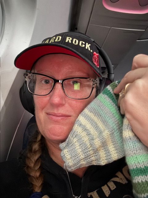 Person wearing a 'Hard Rock' cap and headphones, holding a finished sock while sitting on an airplane.