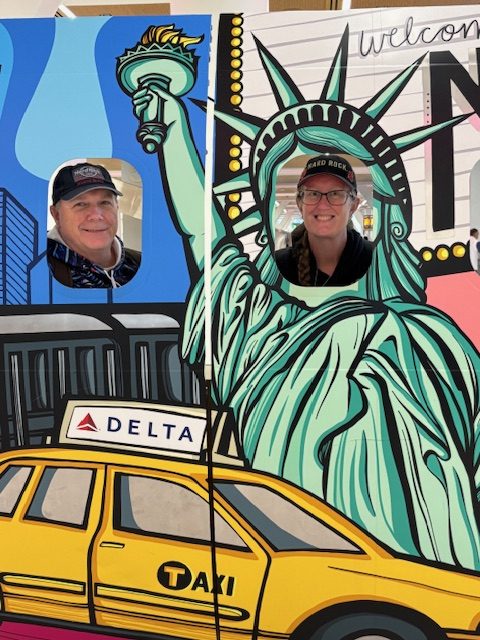 Two people posing behind a colorful cutout featuring the Statue of Liberty and a New York taxi.