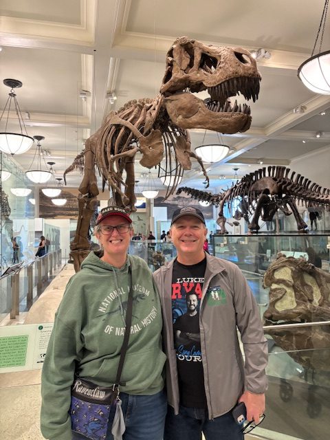 A couple poses in front of a mounted T-Rex dinosaur skeleton at the American Museum of Natural History.