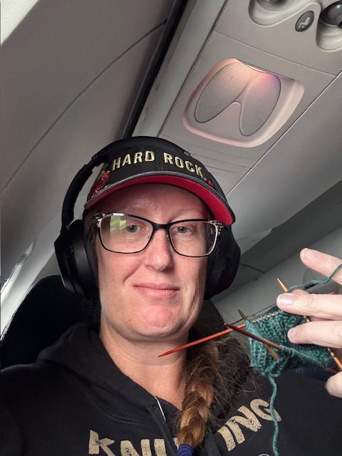 A person wearing a black hoodie and a Hard Rock cap sits on an airplane while knitting.