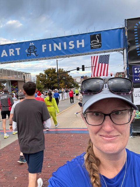 A woman takes a selfie at the start and finish area of a race, with runners in the background and a banner displaying 'START FINISH' above.