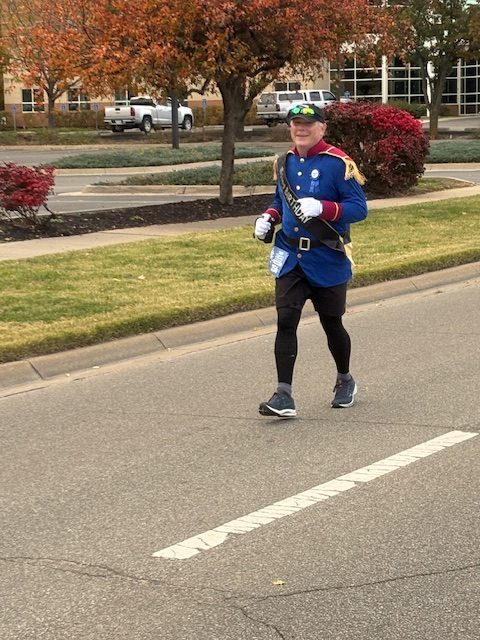 A runner dressed in a colorful costume, including a blue jacket and sunglasses, jogging along a road lined with trees and shrubs during a race.