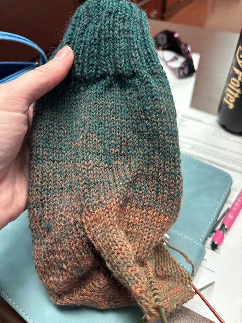 A hand holding a partially knitted travel sock showcasing a color fade from teal to orange, with a work desk and office items in the background.