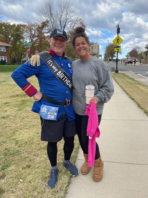 A man wearing a blue jacket with a sash that reads 'It's my BIRTHDAY' stands next to a woman in a gray sweatshirt, both smiling in a park setting during race day.