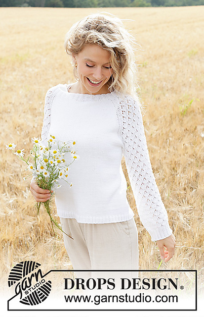A smiling woman wearing a white sweater with lace sleeves, standing in a field of dry grass while holding a bouquet of wildflowers.