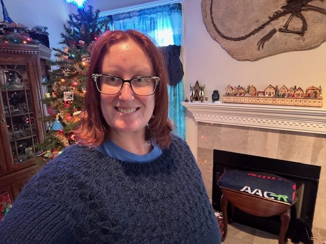 A smiling person wearing a handmade navy sweater stands in a cozy room decorated for the holidays, with a Christmas tree in the background and a fireplace adorned with colorful decorations.