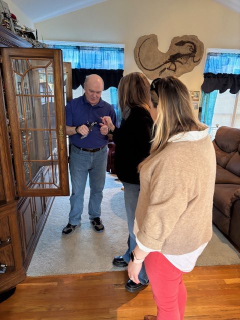 A man in a purple shirt is interacting with two women indoors, with a wooden cabinet in the background. The setting is cozy, with blue curtains and comfortable furniture.