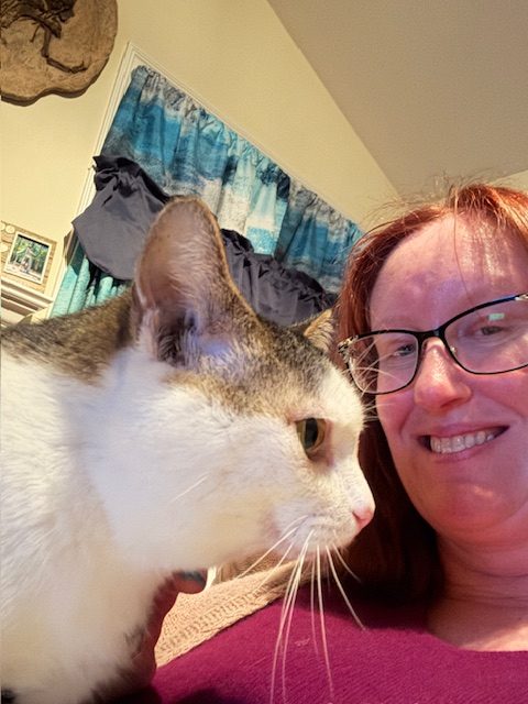 A close-up selfie of a woman with red hair and glasses alongside a white and gray cat. The background features blue curtains and framed photos.