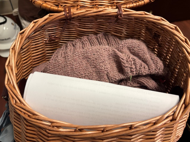 A wicker basket containing a partially completed knitting project in a soft pink yarn, with a sheet of paper partially visible underneath.