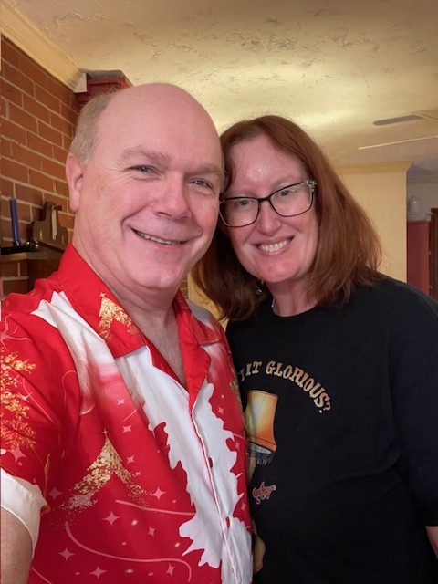 A cheerful selfie of a man wearing a red festive shirt and a woman with glasses, smiling together in a cozy indoor setting.