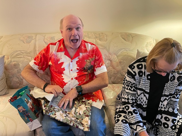A man in a red Hawaiian shirt excitedly unwraps a present while a woman in a patterned black and white sweater sits next to him on a couch.