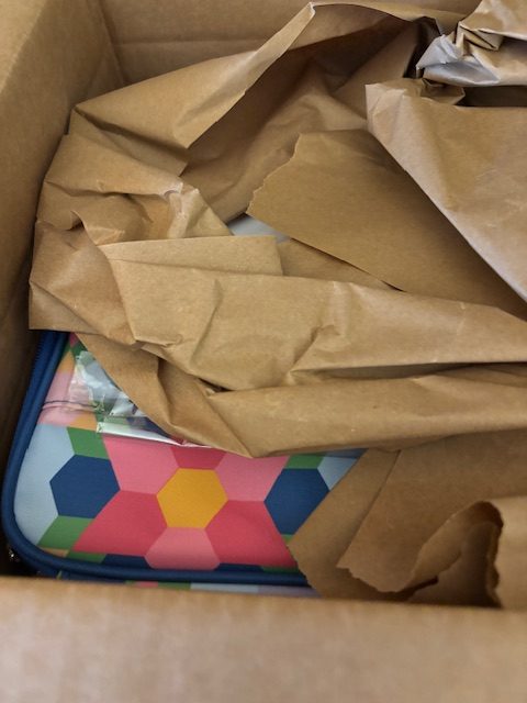 A partially unwrapped colorful item in a cardboard box, surrounded by crumpled brown packing paper.