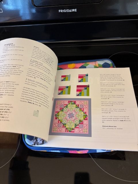 An open quilting instruction book displaying colorful block patterns and construction details, resting on a patterned fabric surface near a stove.