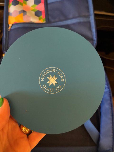 A circular teal fabric cutting mat with the logo 'Missouri Star Quilt Co.' embossed in gold at the center, held in a person's hand.