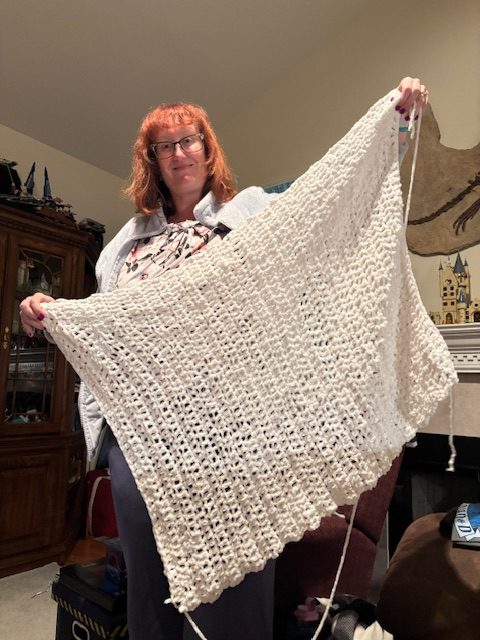 A person with shoulder-length red hair wearing glasses holds up a large white crocheted item, showcasing its intricate pattern, in a cozy indoor setting.
