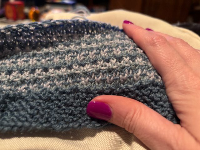 A close-up of a hand with purple nail polish gently touching a knitted fabric in shades of blue and white.