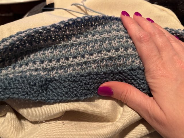 Close-up of a hand with purple nail polish resting on a textured blue knitting project, with a beige fabric underneath.