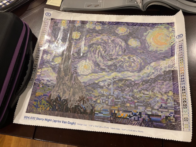 A diamond painting kit featuring a colorful interpretation of Vincent van Gogh's 'Starry Night', displayed on a wooden table with a carrying case beside it.