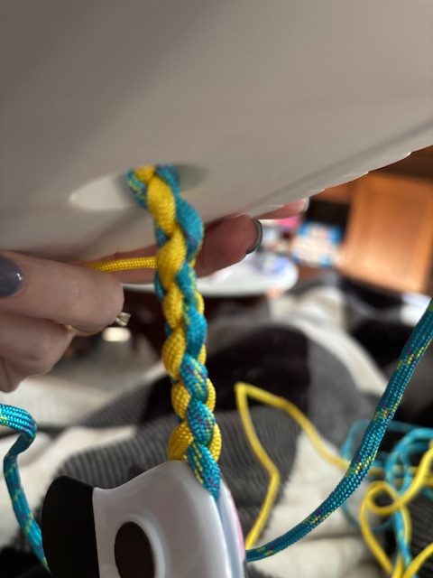 A close-up of a braided cord with blue and yellow strands being held by a hand, with a partially visible white object in the background.