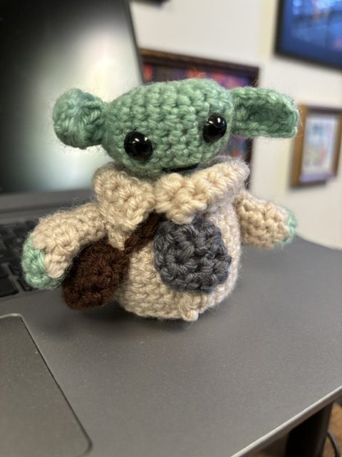 A crocheted plush figure resembling a small alien character with big black eyes, green skin, and large ears, dressed in a beige robe with a brown strap.