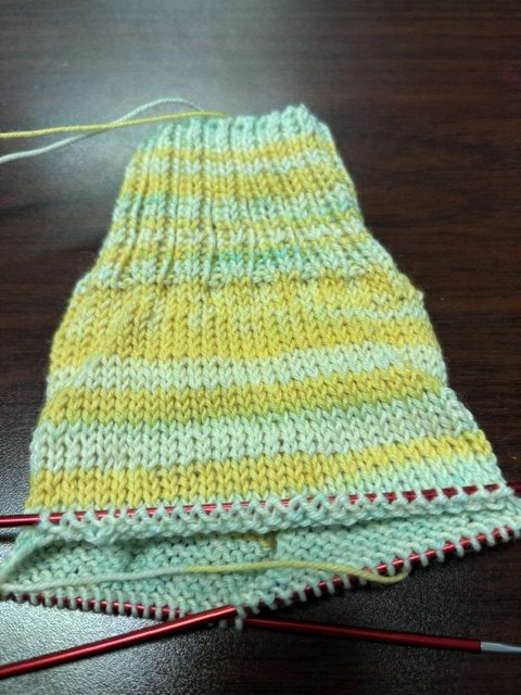 Close-up of a knitting project featuring a striped pattern in yellow and light blue yarn, with knitting needles visible at the bottom.