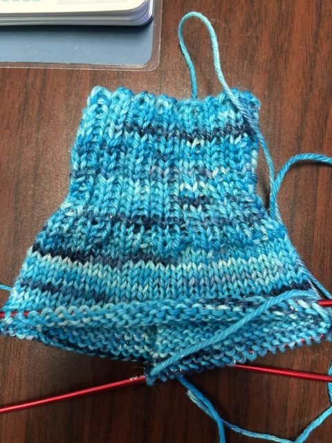 A piece of knitted fabric in various shades of blue, showing ribbed stitch patterns, resting on a wooden table with knitting needles attached.