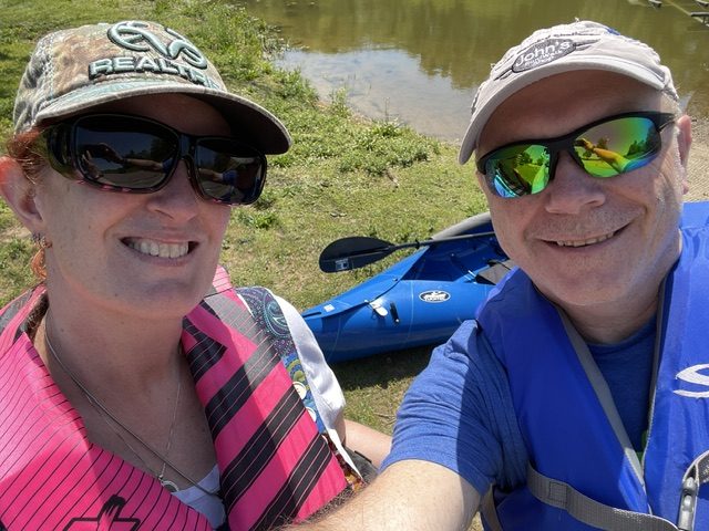 A man and a woman smiling for a selfie while wearing life jackets, with a blue kayak and a pond in the background.