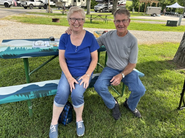 A smiling elderly couple sitting on a picnic bench in a park. The woman, wearing a blue shirt, glasses, and light pants, is sitting beside a man in a gray shirt and blue jeans. They appear to be enjoying a sunny day outdoors.